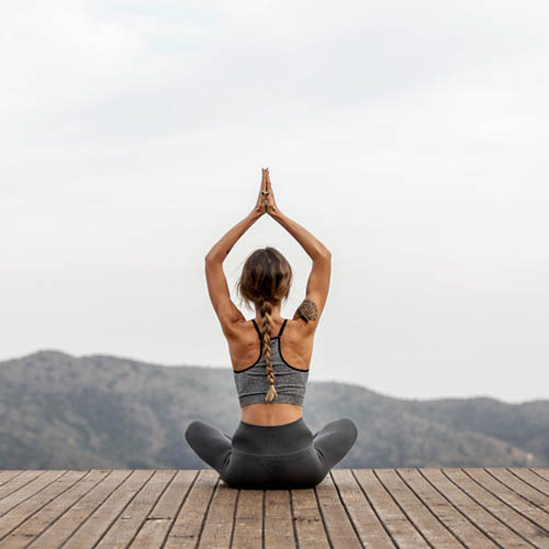 Mulher no exterior, em pose de yoga, de costas para a imagem, com as pernas cruzadas